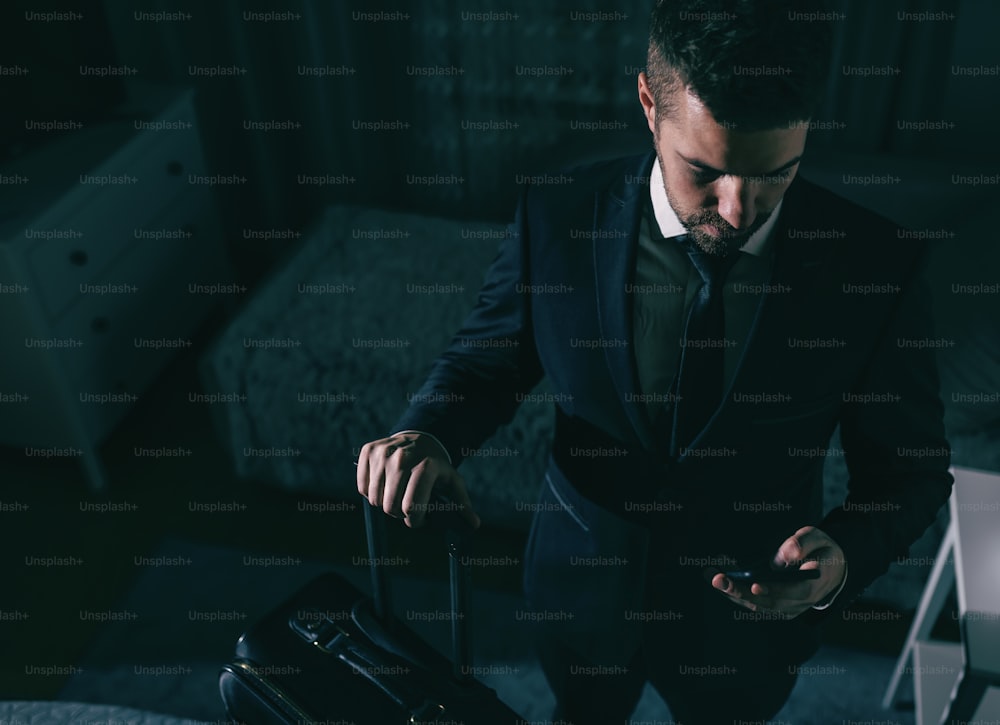 Top view of businessman in suit standing in the bedroom, holding suitcase and using smart phone. Business trip concept.