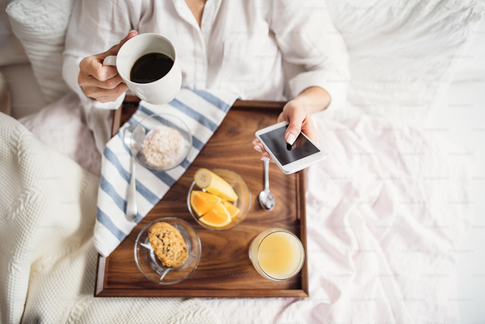 Une section médiane d’une femme allongée dans son lit avec du café et un petit-déjeuner à l’intérieur le matin dans une chambre, à l’aide d’un smartphone. Vue de dessus.