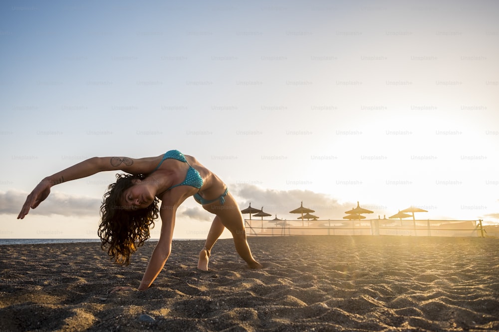 Yoga and pilates fitness workout at the beach for attractive brunette doing exercises - golden sunset light in background - healthy lifestyle for millennial young people - outdoor fit life concept
