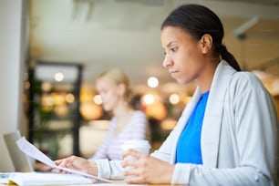 Young serious mixed-race female economist reading paper while sitting in cafe and having drink