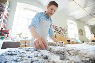 Young craftsman in apron leaning over board while laying mosaic picture in his workshop