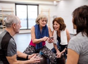 Eine Gruppe von Senioren mit Smartphone im Fitnessstudio, die sich nach dem Training auf fitten Bällen ausruhen.