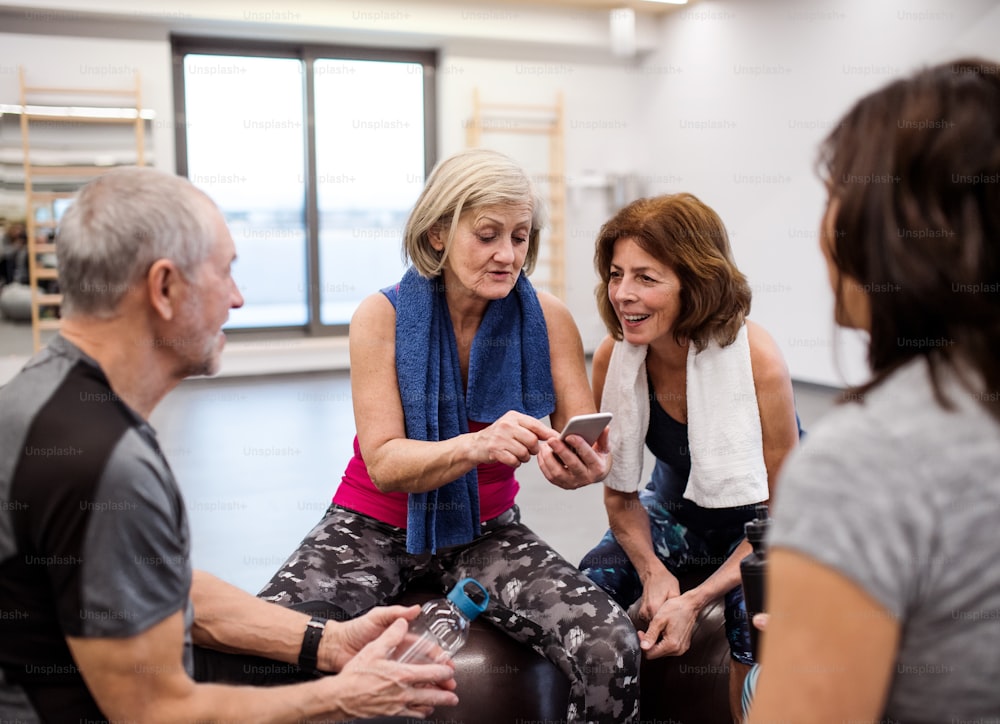 Eine Gruppe von Senioren mit Smartphone im Fitnessstudio, die sich nach dem Training auf fitten Bällen ausruhen.