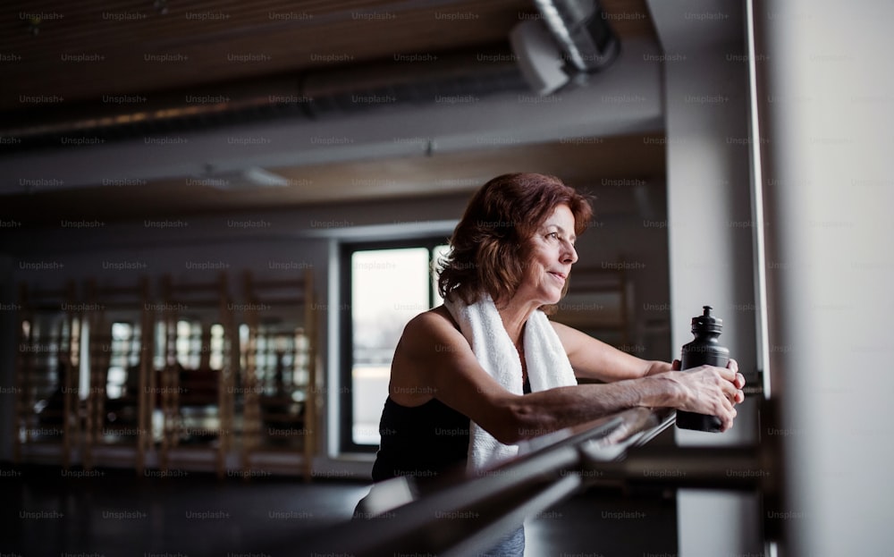 Uma mulher sênior contente com garrafa de água na academia descansando depois de fazer exercícios. Espaço de cópia.