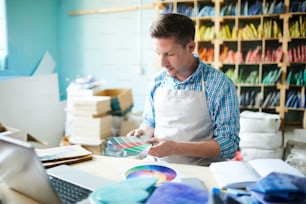 Porträt eines modernen Handwerkers, der die Farbpalette wählt und Muster hält, während er am Tisch in einer modernen Werkstatt sitzt, Kopierraum