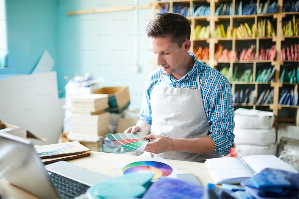 Retrato de un artesano moderno que elige la paleta de colores que sostiene muestras mientras está sentado en la mesa en un taller moderno, espacio de copia