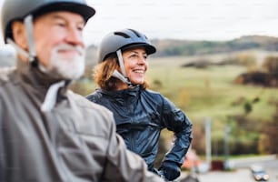 An active senior couple with helmets and electrobikes standing outdoors on a road in nature. Copy space.