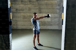 Dedicated muscular man in sportswear doing exercises with kettlebell while standing in gym gym.