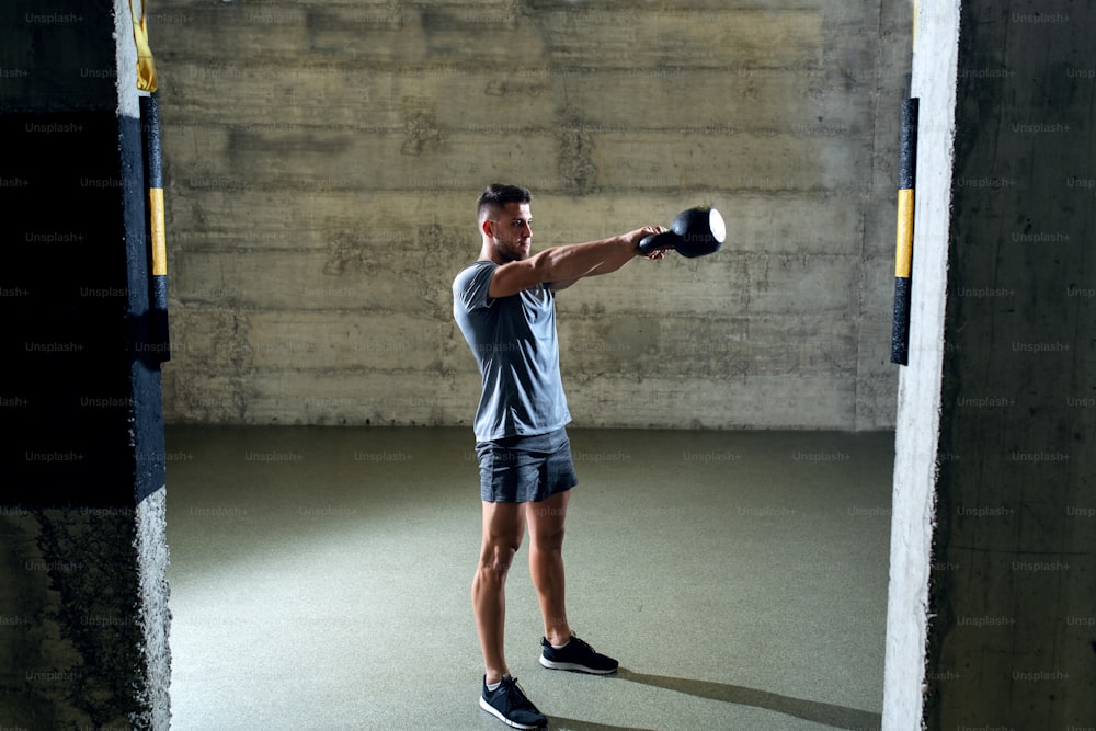 Hombre musculoso dedicado en ropa deportiva haciendo ejercicios con pesas rusas mientras está de pie en el gimnasio.