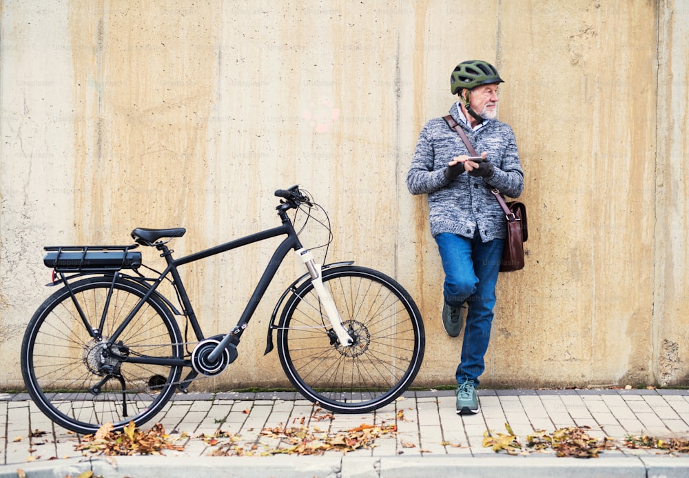 街中で屋外に立ち、コンクリートの壁にもたれかかってスマートフォンを使っている電動自転車を持ったアクティブな年配の男性。スペースをコピーします。