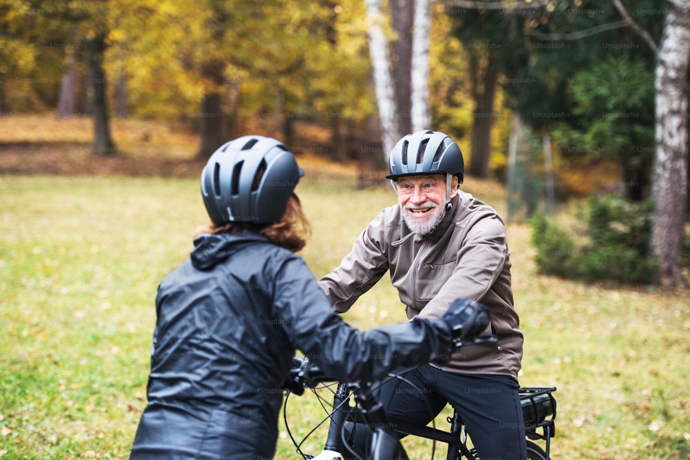 Un couple de personnes âgées actives avec des casques et des vélos électriques debout à l’extérieur sur une route dans la nature, se regardant.