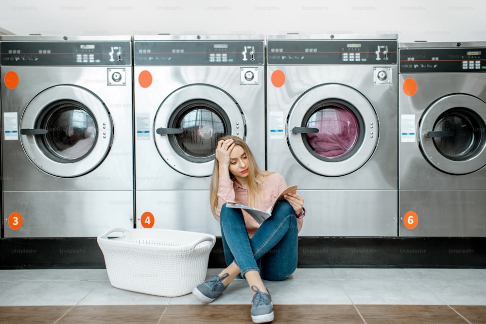 Young and sad woman waiting for the clothes to be washed sitting on the floor at the self-service laundry