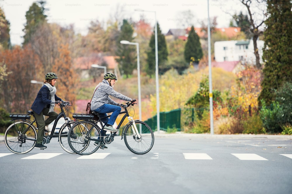 Couple de personnes âgées actives avec des vélos électriques à l’extérieur traversant une route en ville. Espace de copie.