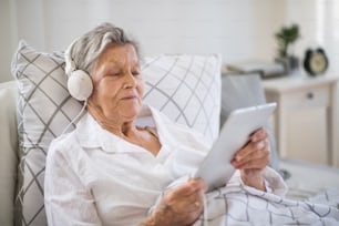 A sick senior woman with headphones and tablet lying in bed at home or in hospital, listening to music.