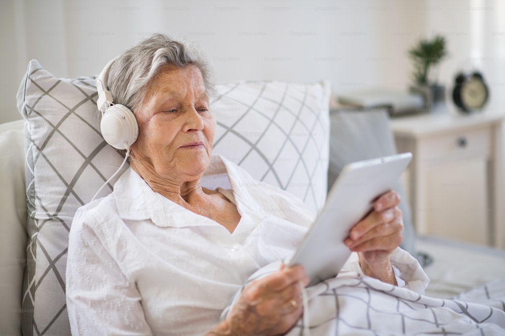 Une femme âgée malade avec des écouteurs et une tablette, allongée dans son lit à la maison ou à l’hôpital, écoutant de la musique.