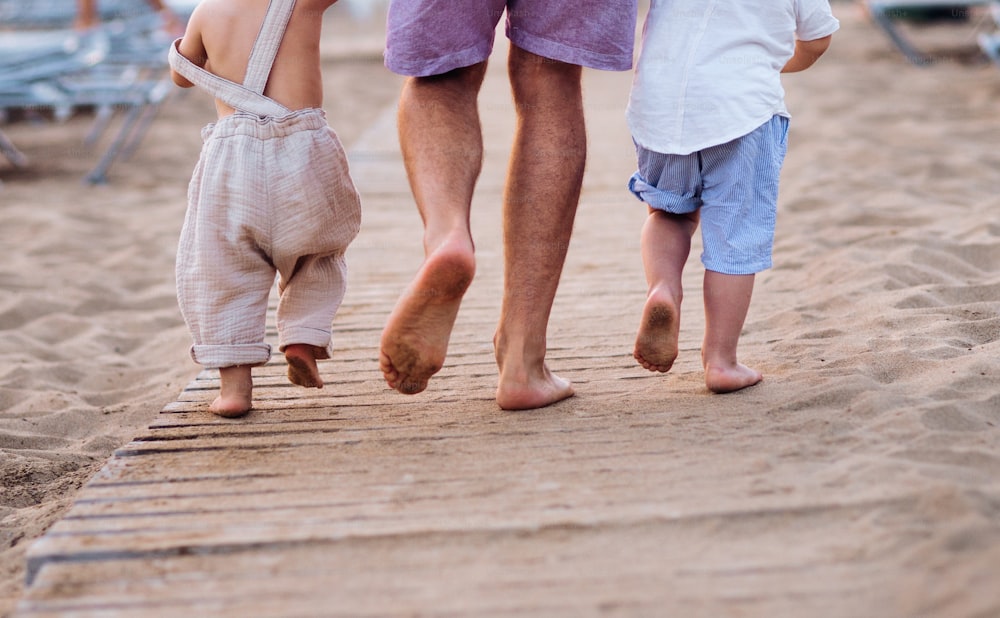 Una sección media de padre con dos niños pequeños caminando descalzos en la playa de arena en vacaciones de verano. Una vista trasera.