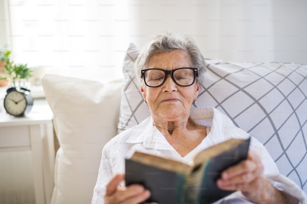 안경을 쓴 아픈 노인 여성이 집이나 병원에서 침대에 누워 성경을 읽고 있습니다.