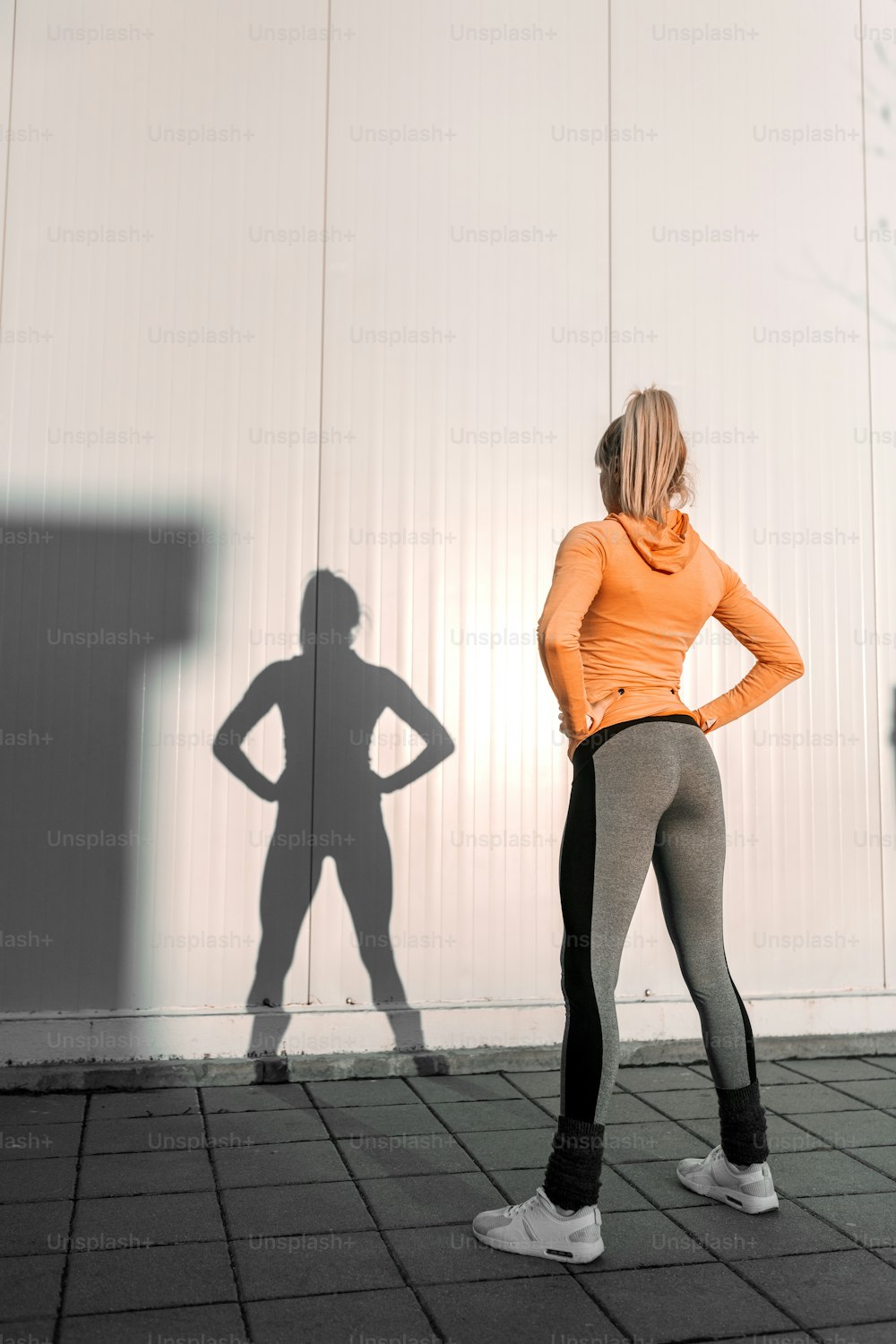 Slim sporty blonde girl in sportswear standing in front of white background and holding hands on hips. Backs turned.