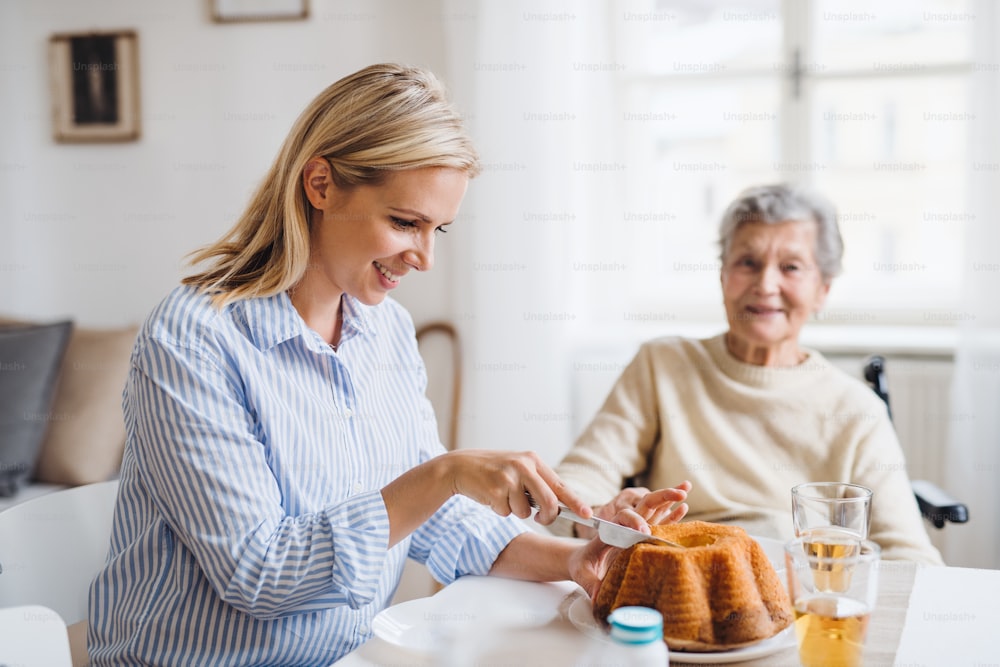 Uma mulher idosa em cadeira de rodas com um visitante de saúde sentado à mesa em casa, cortando um bolo.