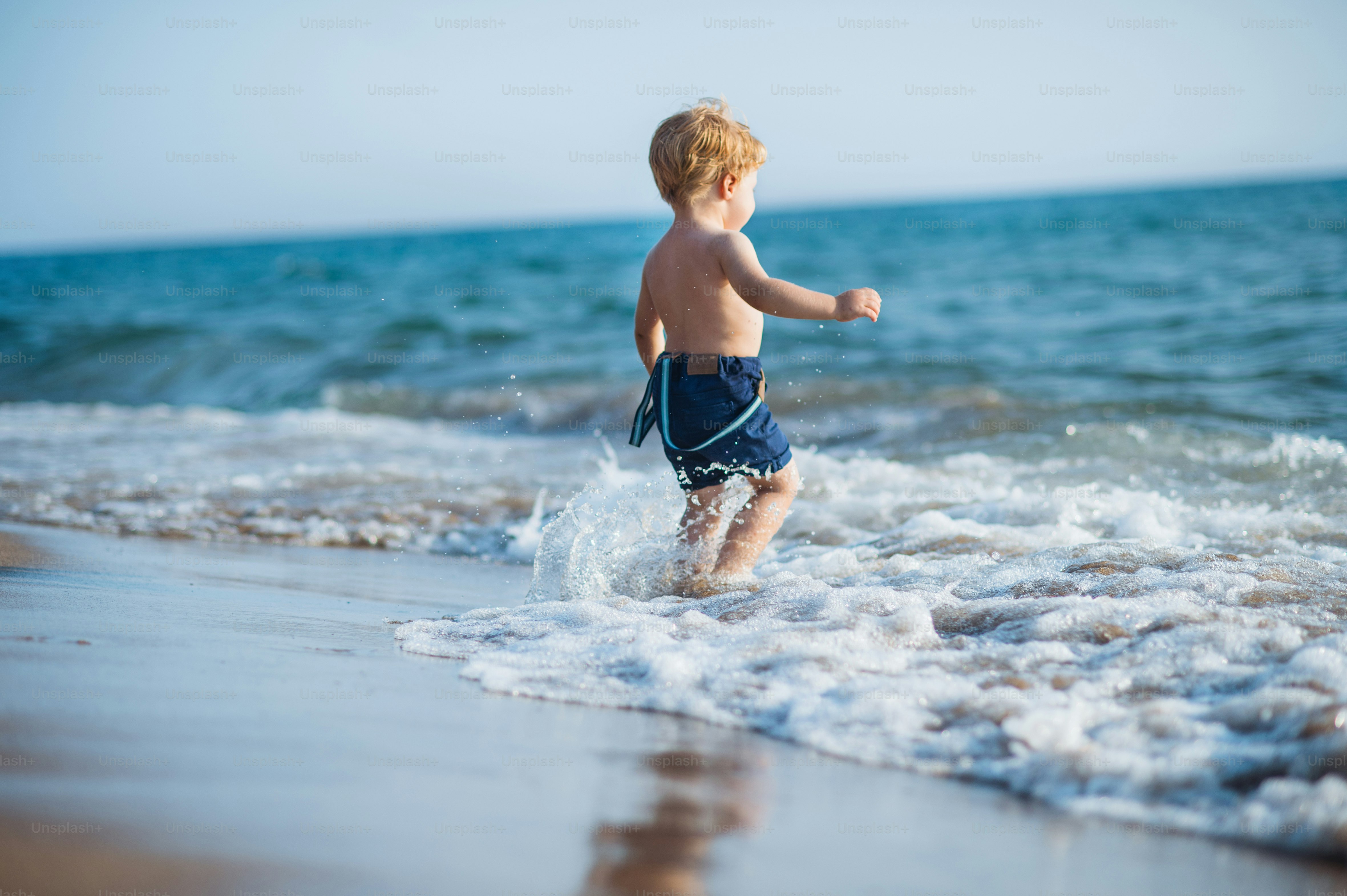 Seorang bocah laki-laki kecil yang lucu dengan celana pendek berjalan di air di pantai pada liburan musim panas.
