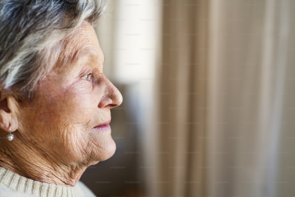 Eine Nahaufnahme des Porträts einer älteren Frau zu Hause, die aus einem Fenster schaut.