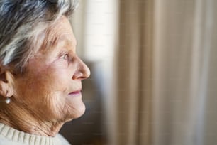 Un ritratto ravvicinato di una donna anziana a casa, che guarda fuori da una finestra.