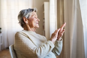A portrait of a senior woman sitting on a chair at home, looking out of a window.