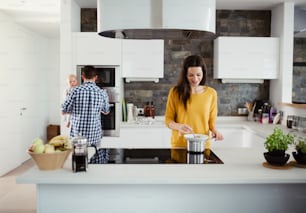 Un ritratto di giovane famiglia in piedi in una cucina di casa, un uomo che tiene in braccio un bambino e una donna che cucina.