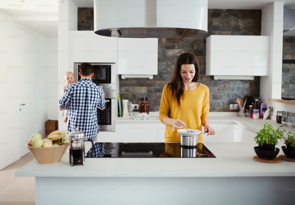 Un retrato de una familia joven de pie en una cocina en casa, un hombre sosteniendo a un bebé y una mujer cocinando.