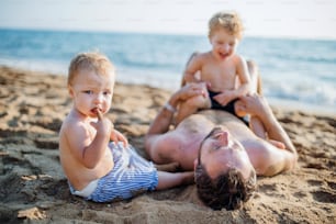 A father with two toddler children lying on sand beach on summer holiday, playing.