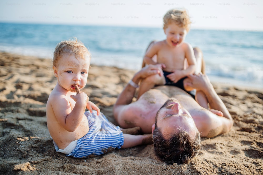 여름 휴가에 모래 해변에 누워 놀고 있는 두 명의 유아 자녀를 둔 아버지.