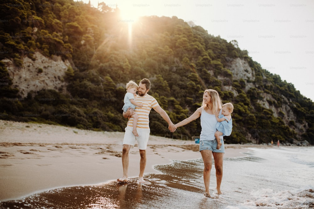 A family with two toddler children walking on beach on summer holiday at sunset. A father and mother carrying son and daughter in the arms, holding hands.