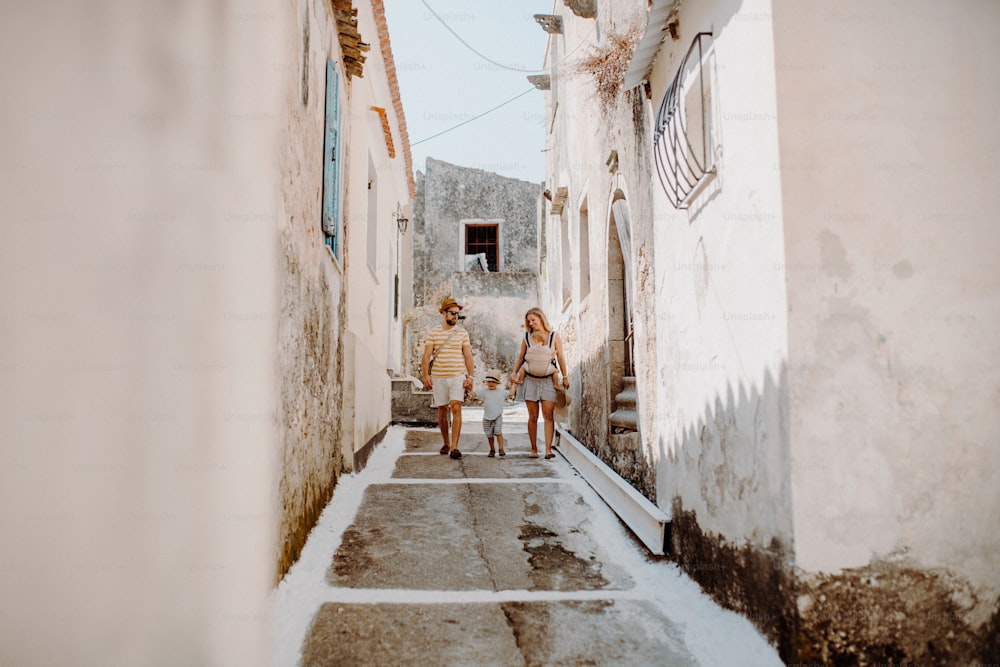 A family with two toddler children walking in town on summer holiday. A father and mother with son and daughter in baby carrier on a narrow street.
