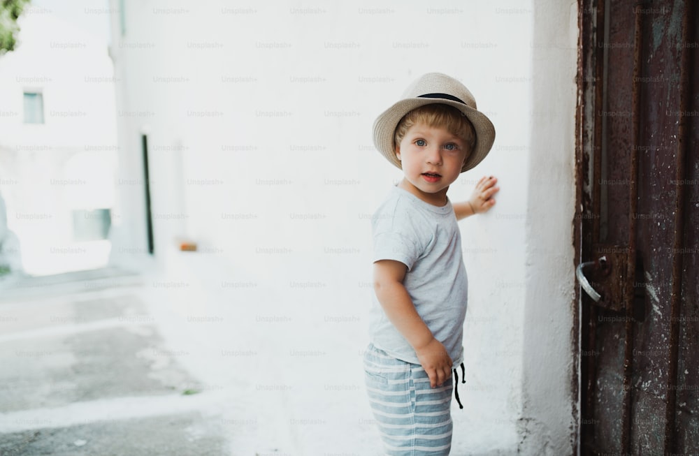 A small toddler boy with hat standing by a house in town on summer holiday. Copy space.