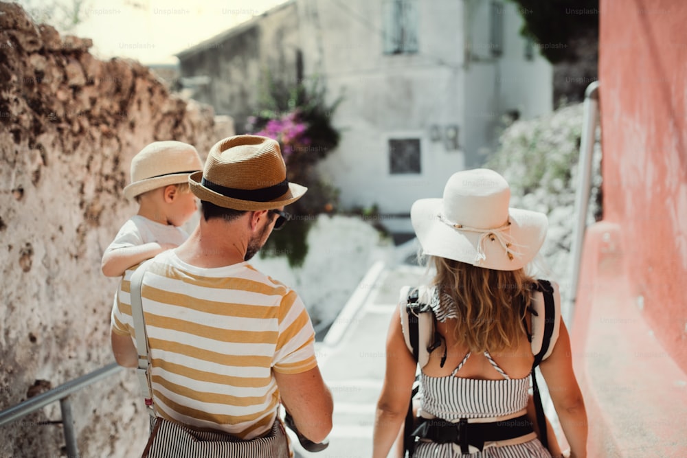 Una vista posteriore di una famiglia con due bambini piccoli che camminano in città durante le vacanze estive. Un padre e una madre che portano in braccio un figlio e una figlia.