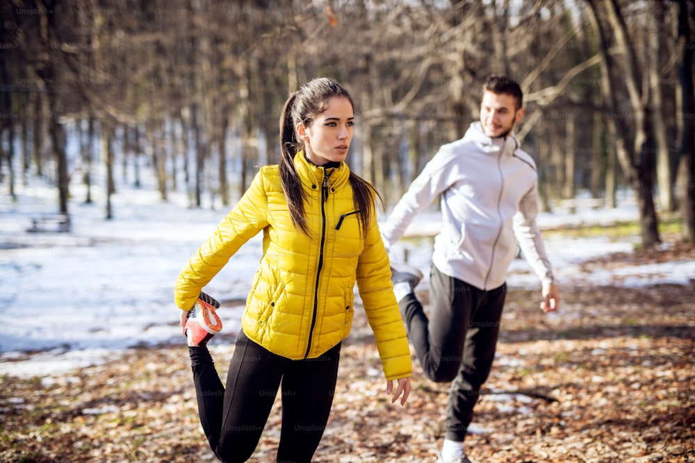 Portrait view of focused handsome hardworking sporty fitness girl in winter sportswear stretching outside with personal trainer in nature.