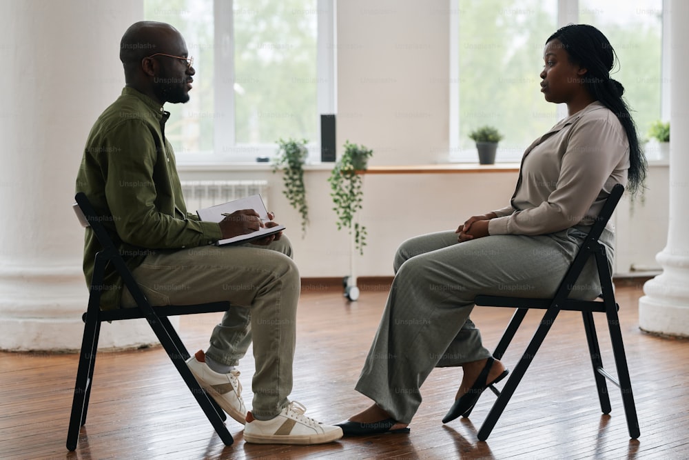Vista lateral do jovem psicólogo afro-americano do sexo masculino com caderno consultando a paciente do sexo feminino sentada na frente dele na sessão