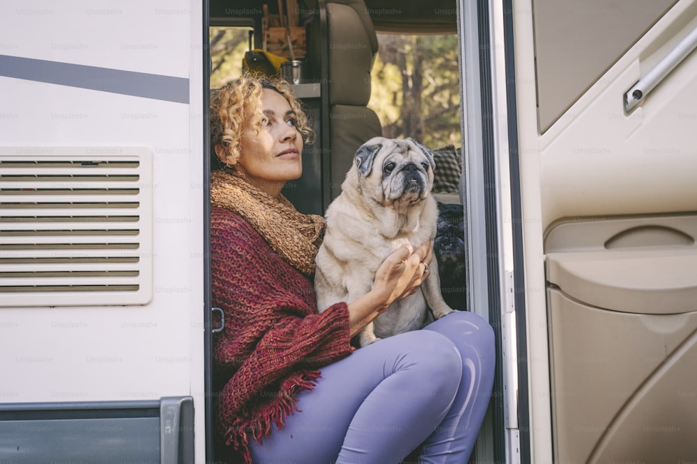 One woman sitting on the camper van door with old nice pug dog looking outside and enjoying relax and freedom. Female people living on a rv motorhome with animals and travel the world. Best friend