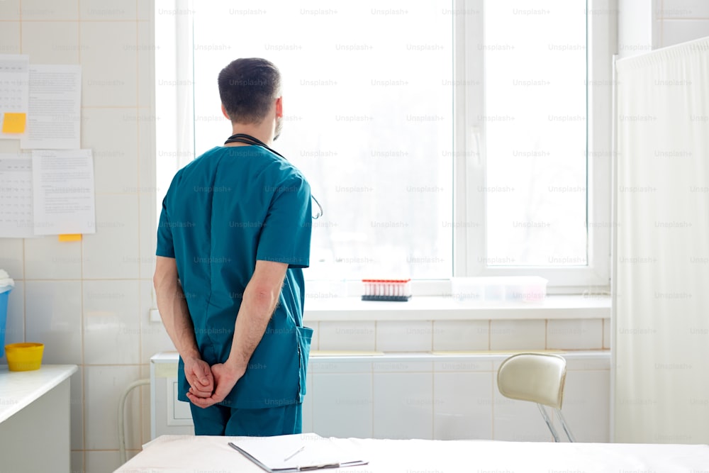 Vista posterior de un clínico contemporáneo en uniforme mirando a través de la ventana en el hospital