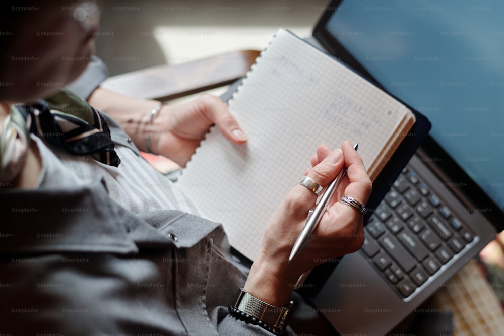Hand einer Ökonomin oder Freiberuflerin mit Stift über Seite des Lehrbuchs und Laptop-Tastatur, die finanzielle Notizen macht, während sie den Plan aufschreibt
