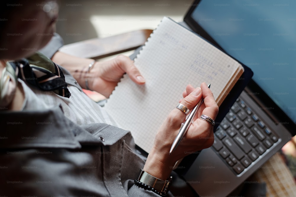 Hand of female economist or freelancer with pen over page of copybook and laptop keyboard making financial notes while writing down plan