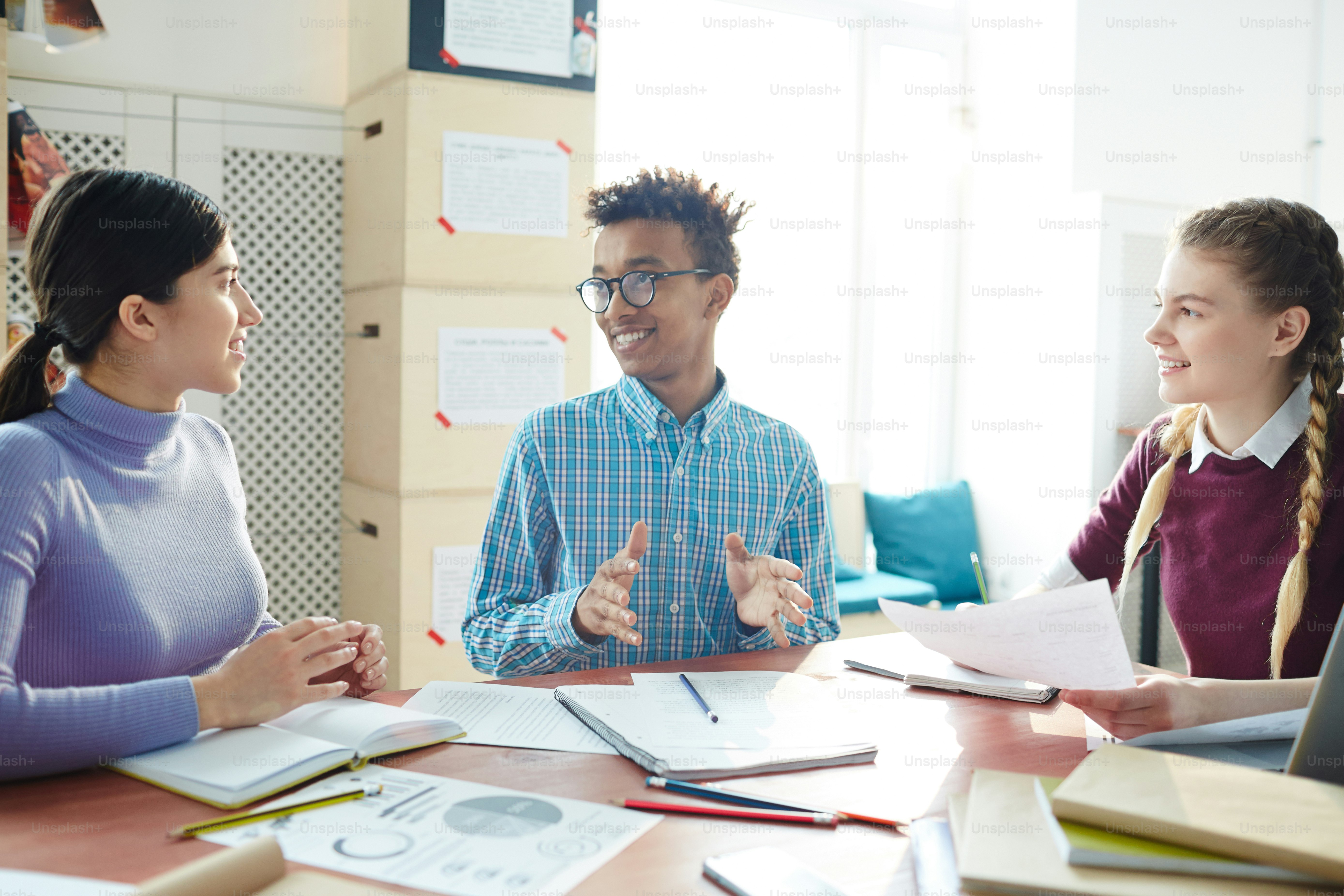 Young guy and two girls in casualwear sitting by desk and preparing for college project after classes