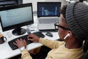 Sobre el ángulo de un joven negro en ropa informal escuchando música con auriculares mientras decodifica datos frente a la pantalla de la computadora