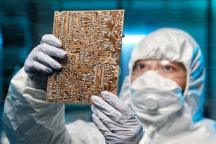 Gloved female specialist in protective coveralls and respirator holding circuit board in front of herself while working in laboratory