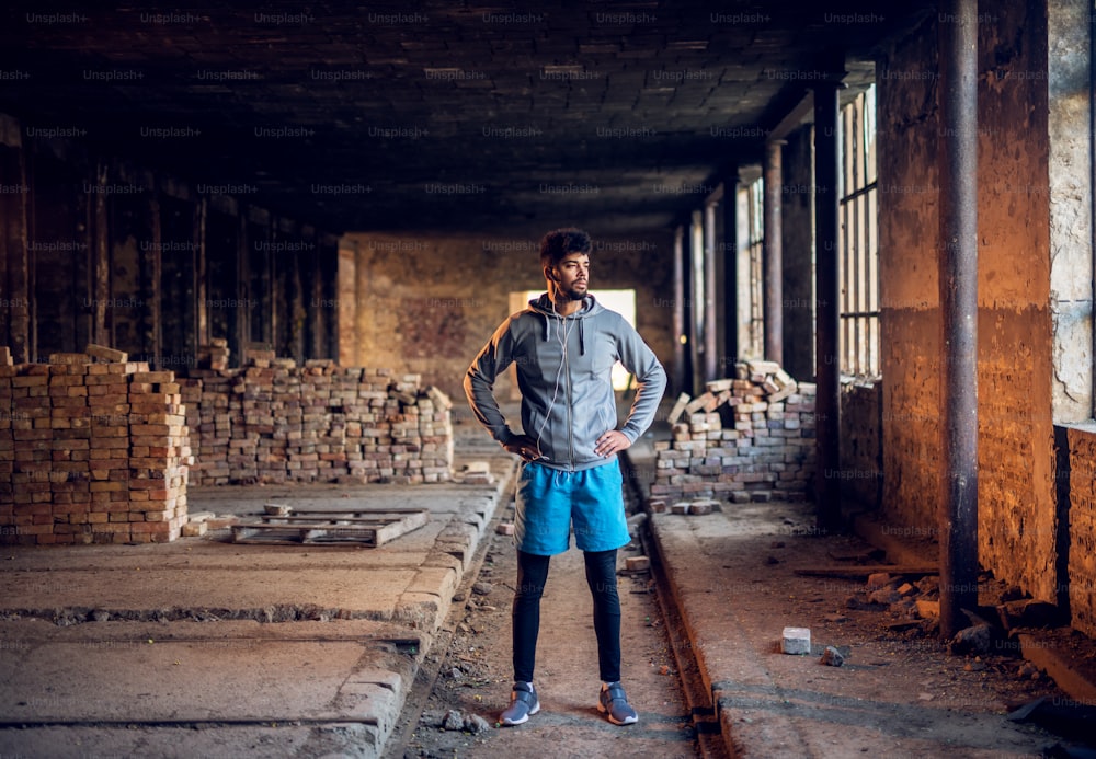 Jeune coureur afro-américain sportif se reposant avec des écouteurs dans un endroit abandonné et regardant au loin.