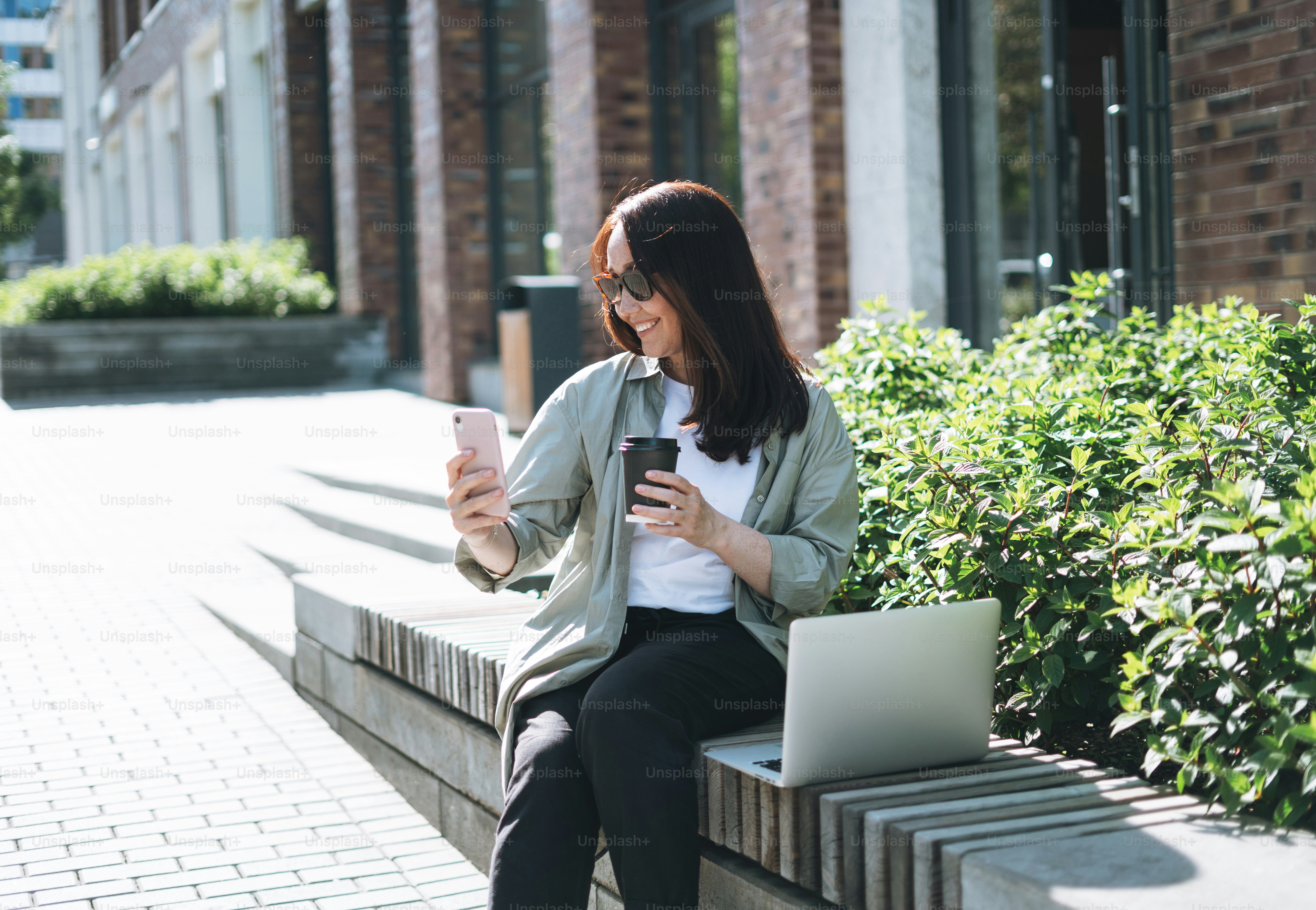 Adult smiling brunette business woman forty years in stylish shirt working on laptop and using mobile phone at public place on bench at city street