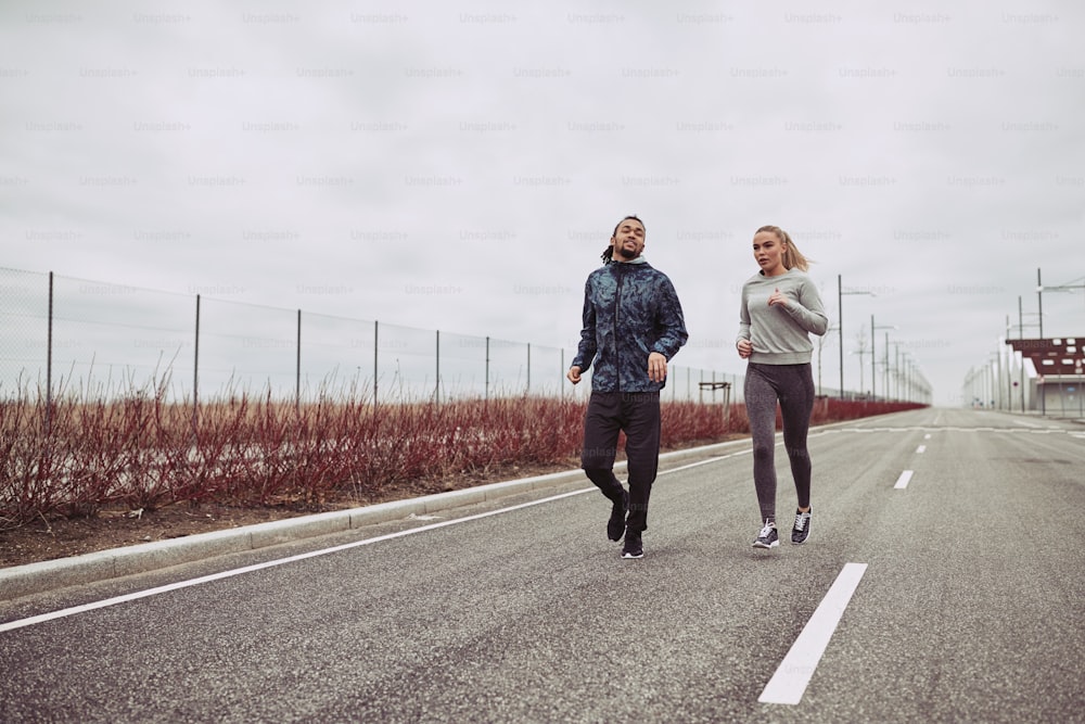 Diverse young couple in sportswear looking focused while jogging together along a road in the country