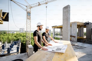 Two workers in black t-shirts and protective harhats working with drawings at the construction site outdoors