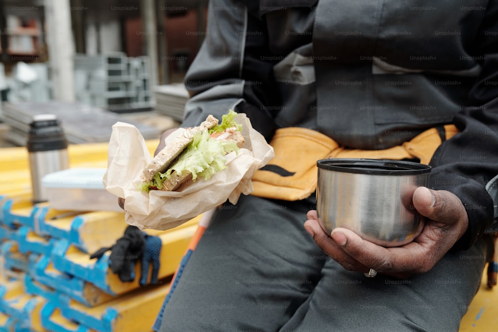 Mano dell'operaio afroamericano del cantiere che tiene la tazza con il tè caldo ed il panino mentre pranza sul posto di lavoro alla pausa