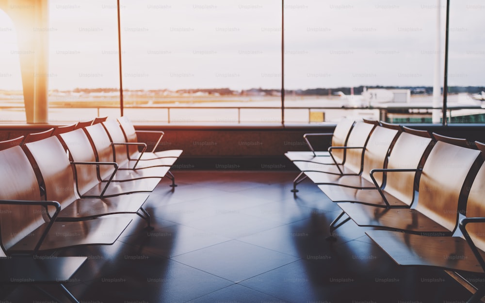 Two empty rows of wooden armchairs in front of each other in waiting room of departure area indoors of modern airport terminal, huge window and take-off field behind in defocused background